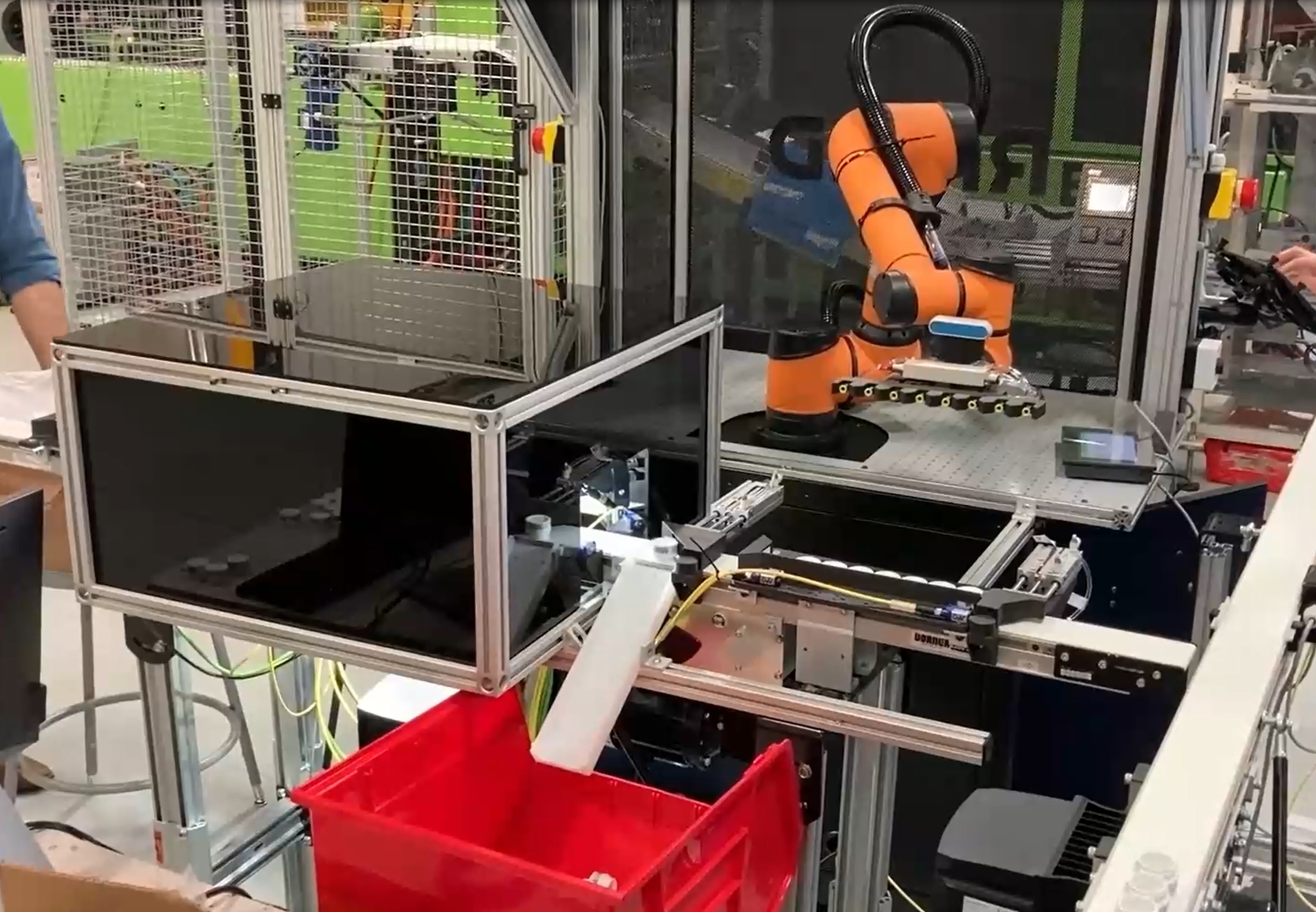 An orange robot with a horizontally oriented suction gripper waits to pick up product next to a polarized box where small jars are inspected by a camera system.