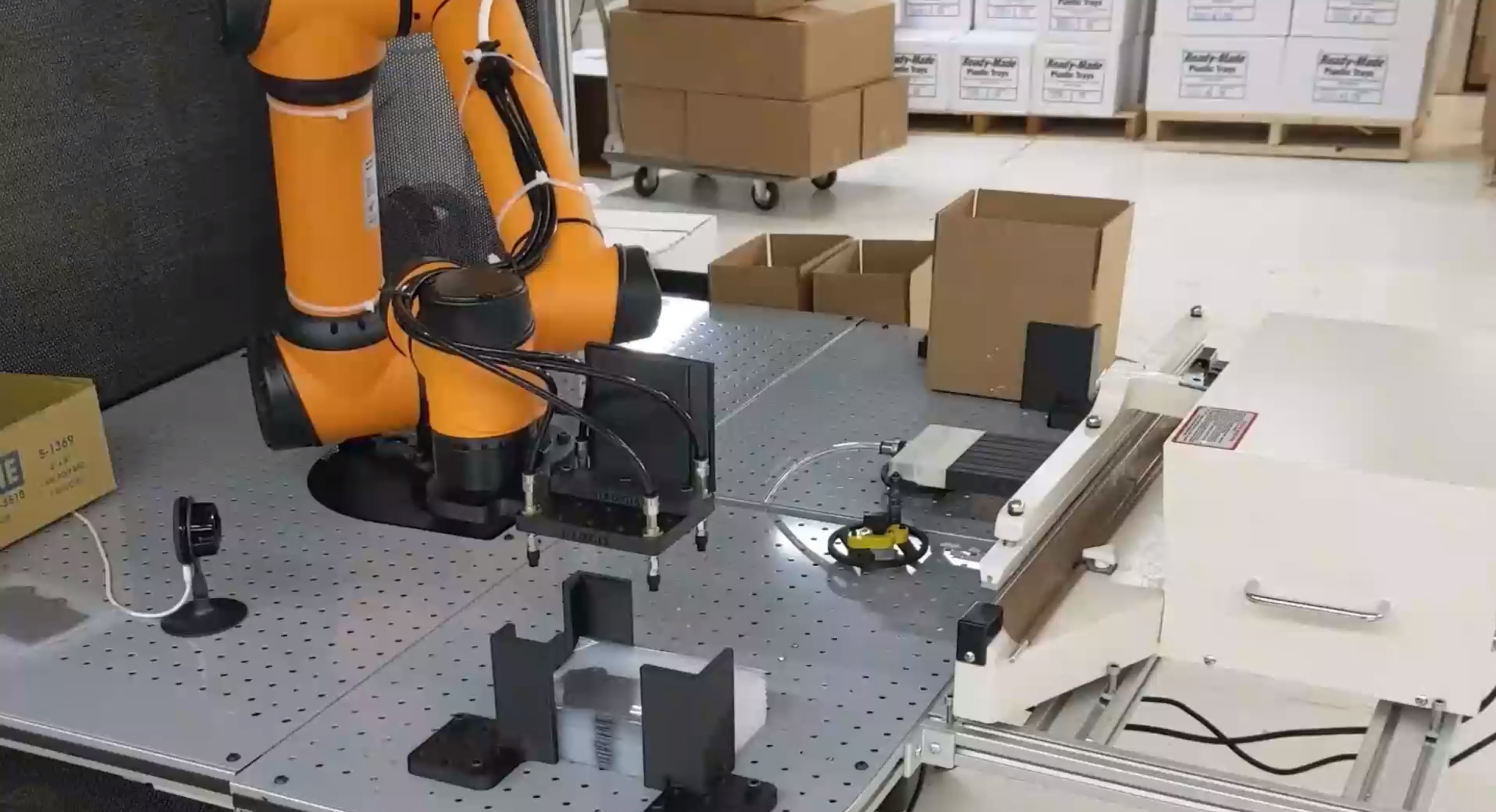 An orange cobot with a custom gripper prepares to pick up a fragile graphite part in a poly bag.