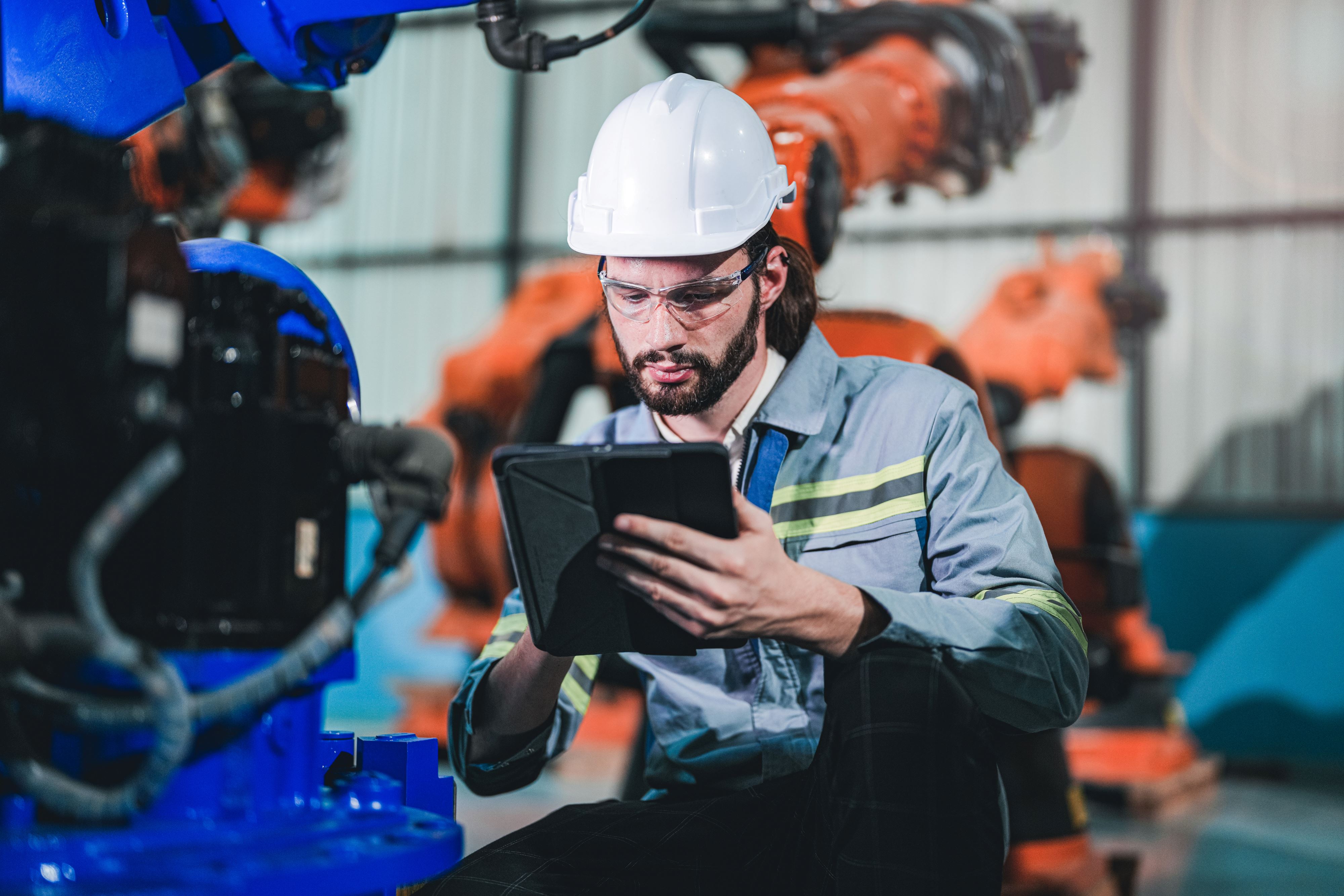 A white man with a beard and long hair, wearing a hard hat and working on a tablet, sits near a pair of large industrial robotic arms.