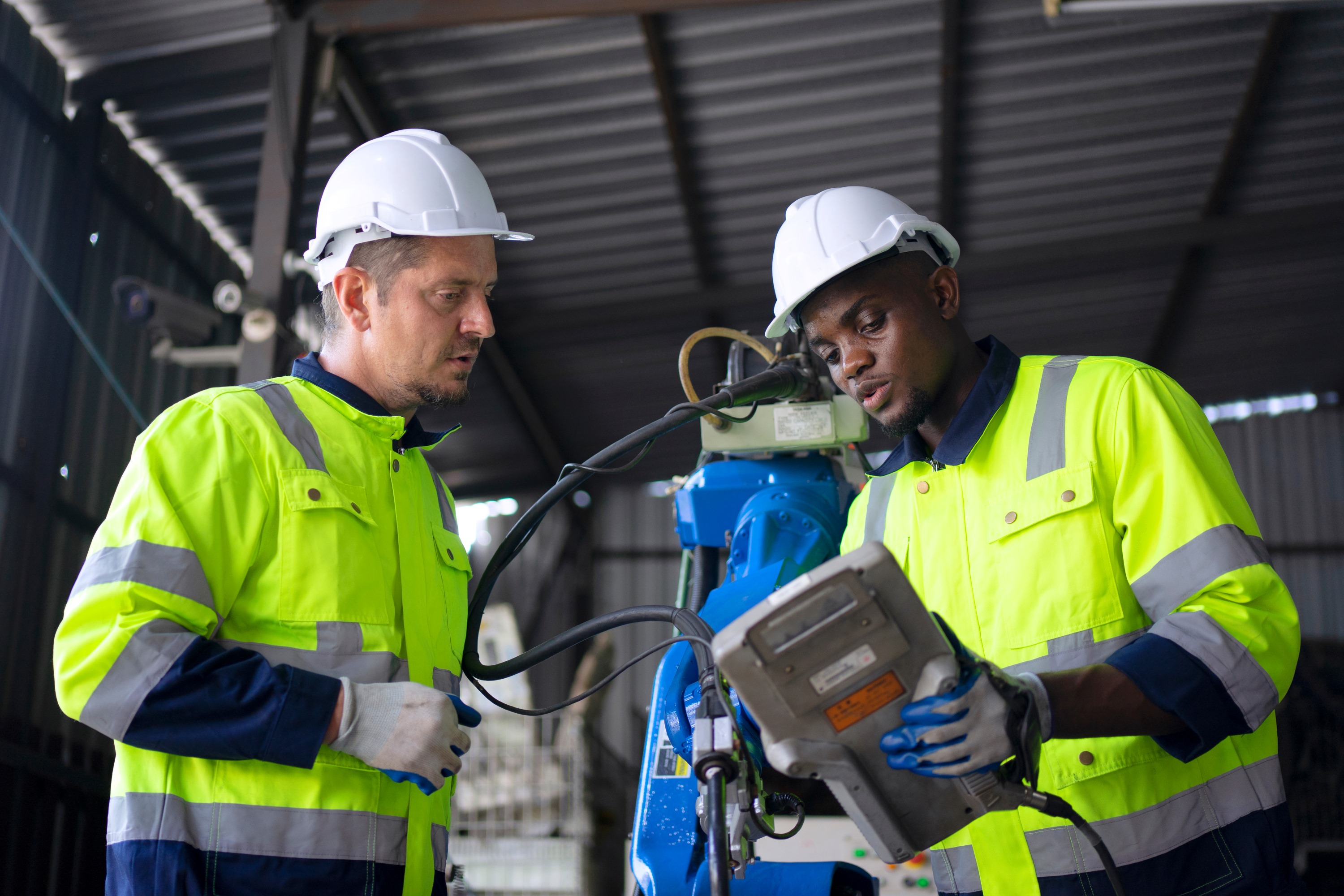 A Black man and a white man wearing high visibility jackets and hard hats work together to maintain an industrial robot. The Black man is holding the robot's teach pendant, and showing it to the white man.