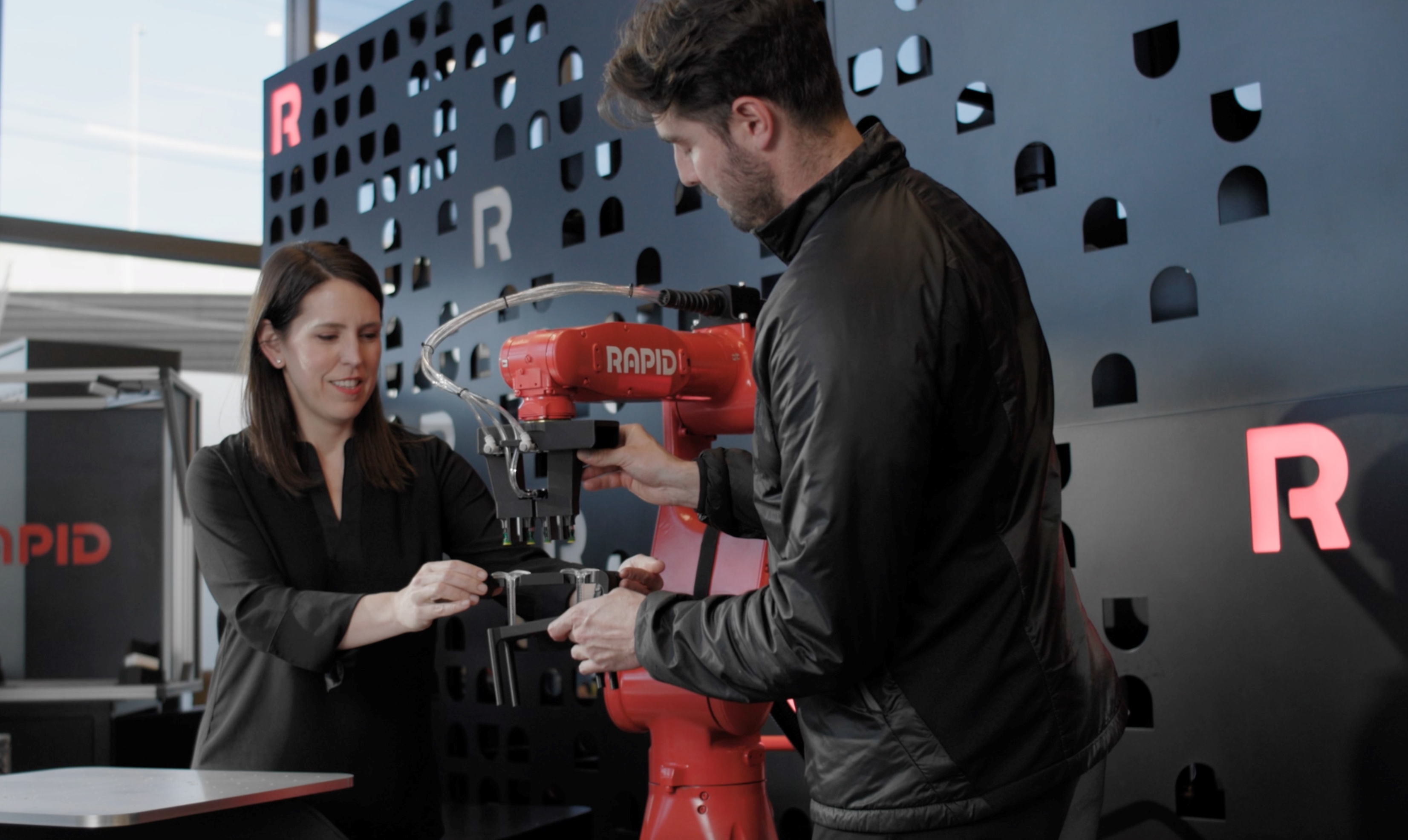 A white woman and a white man both wearing black outfits work together to test a Yaskawa industrial arm with the Rapid logo painted on the side
