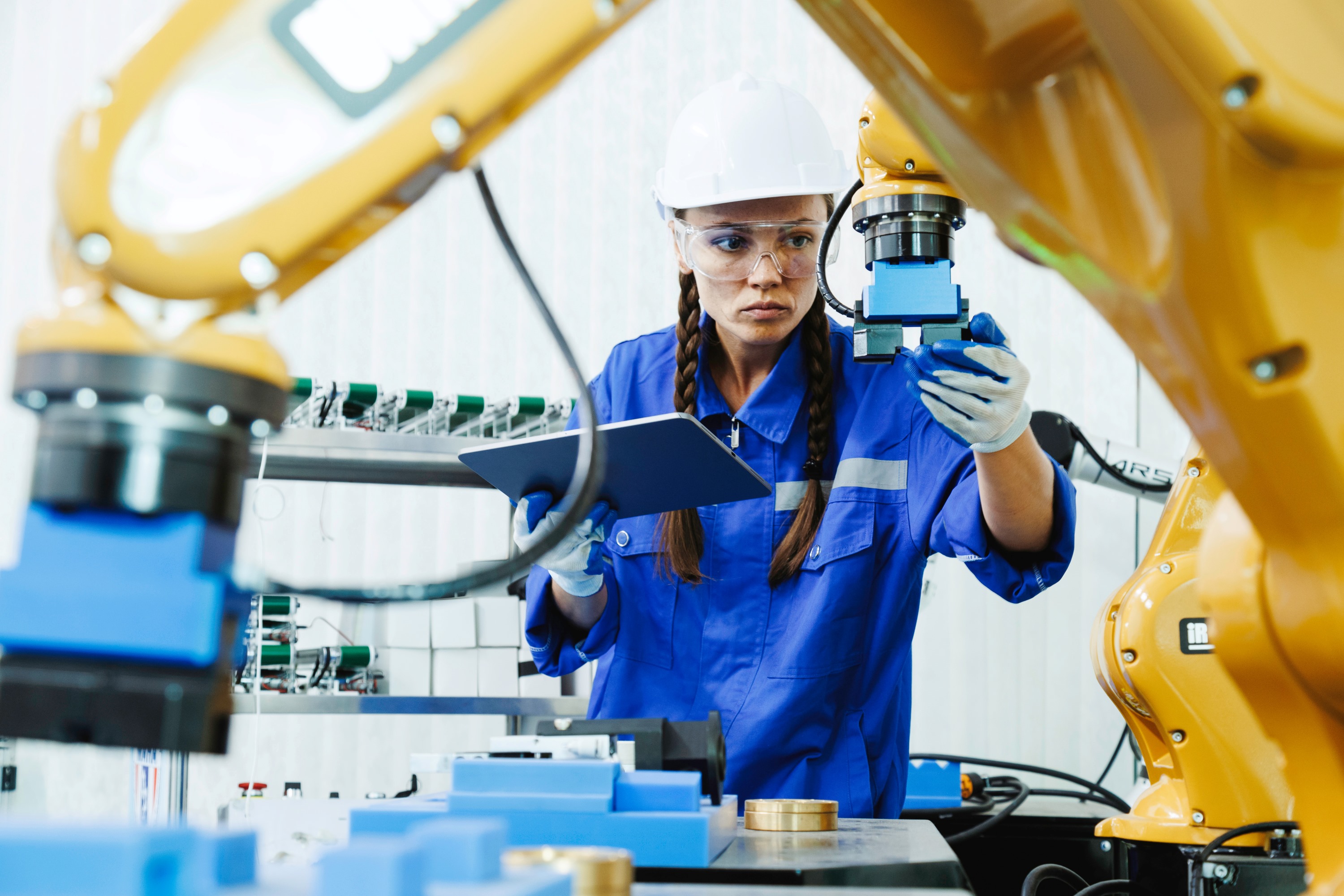 A woman with her hair in braids, wearing work clothes and a hard hat, checks gripper placement on an industrial robot. She is holding a tablet HMI in her other hand.