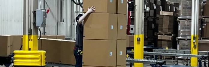 A worker lifts a large, heavy box over their head onto the top of a palletized stack.
