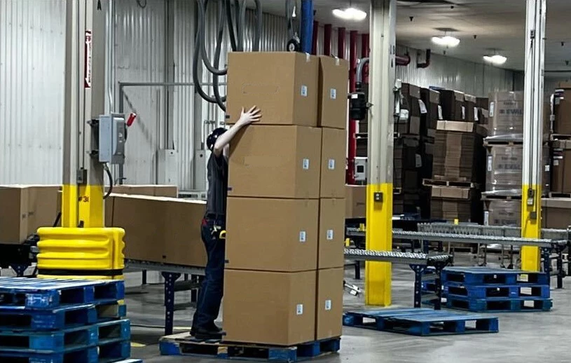 A worker lifts a large, heavy box over their head onto the top of a palletized stack.