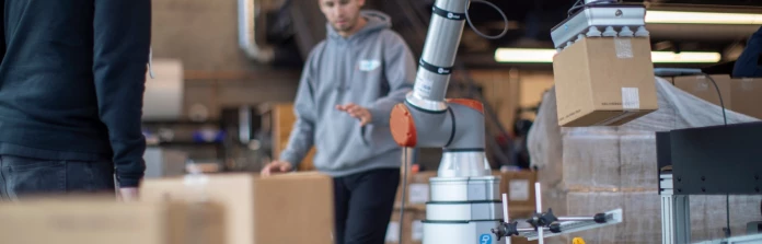 Two workers in hoodies near a UR5 cobot on an Onrobot 7th axis palletizing medium-sized boxes coming off a roller belt.