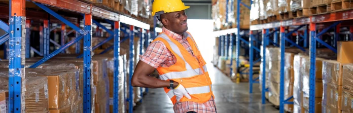 A Black man in a hard hat, safety vest, and work gloves stands in a warehouse full of plastic wrapped pallets and stretches his back