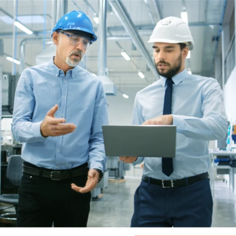 2 men walking through a factory looking at a laptop.