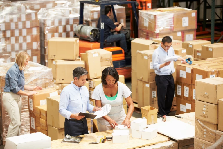 A diverse group of people work in a disorganized warehouse, performing various tasks from driving a forklift to labeling and preparing boxes for shipping.