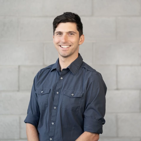 David Hallock wearing a navy button down, smiling in front of a light colored cinderblock wall.