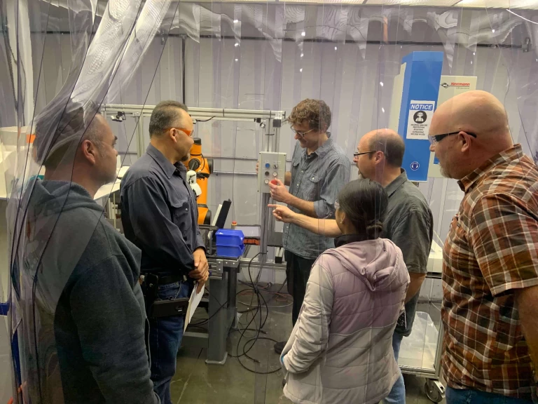 5 people are surrounding a man demonstrating and explaining how the Rapid Machine Operator works. The explainer has his right hand wrapped around the buttons that are attached on a workcell and his left hand pointing onto a button and explaining the function of the button.