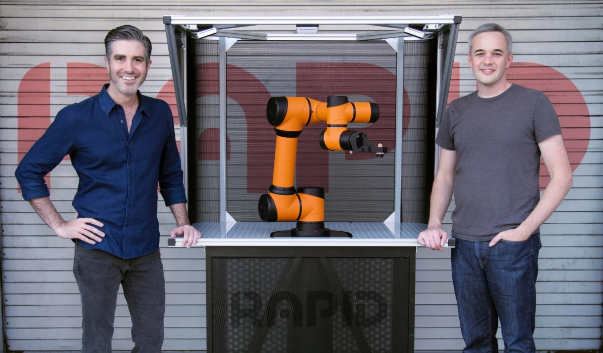 Jordan Kretchmer ( CEO of Rapid Robotics) Leaning with his left hand on the right side of a  workcell of an RMO and wearing a blue button down shirt with gray jeans. Lawrence Ruddick ( CTO of Rapid Robotics) is standing on the right side of the RMO with his right hand leaning on the workcell wearing a gray plain shirt and blue Jeans.